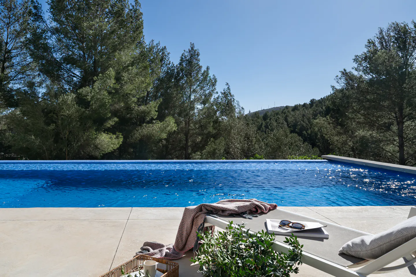 Outdoor pool with blue water, lounge chair with towel, book, and sunglasses. Trees and blue sky in the background.
