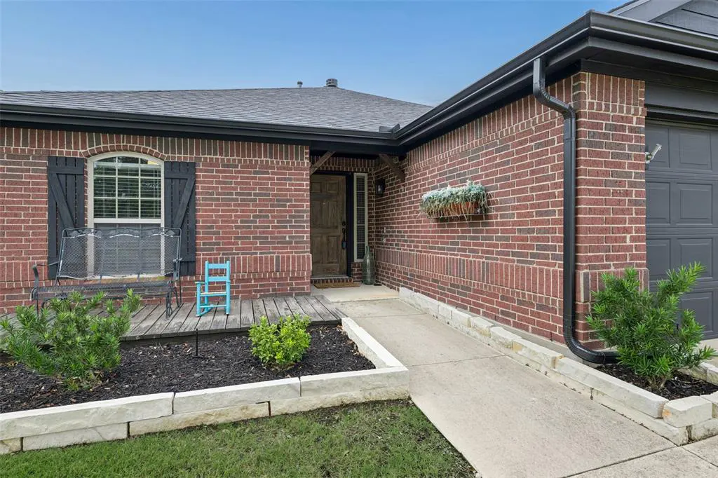 Brick house with a gray roof, black shutters, and a wooden porch. A blue child's chair sits next to a metal bench. A concrete ramp leads to the front door.