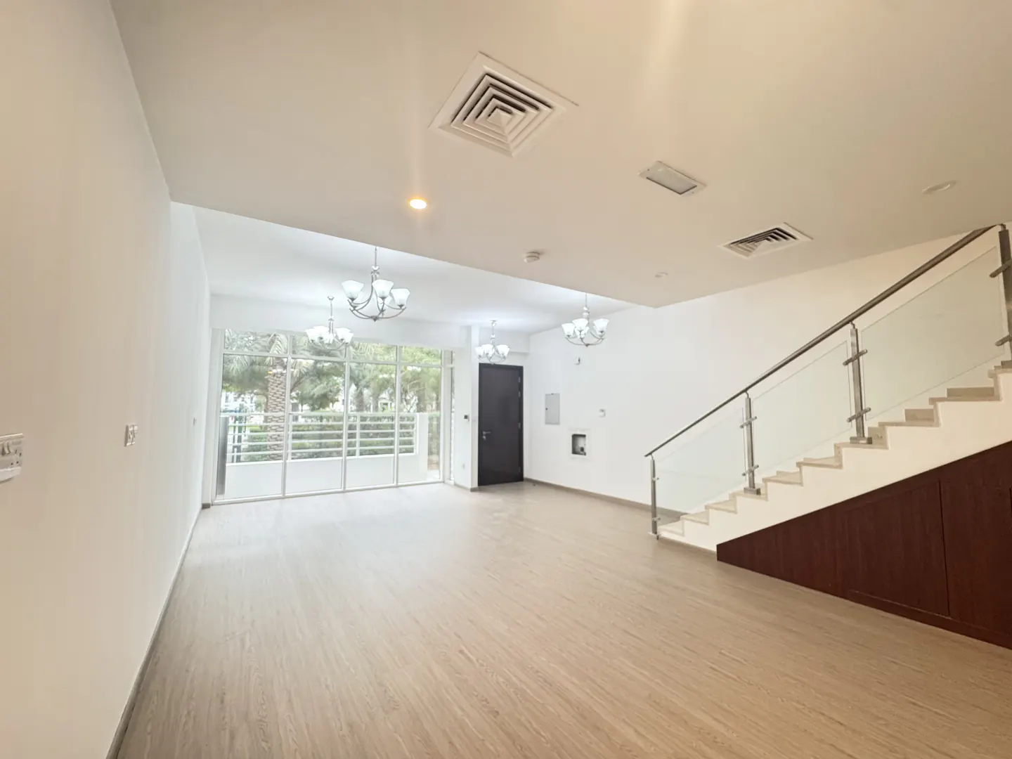 Bright, empty living room with wood floors, white walls, and a staircase with glass railing. Chandeliers hang from the ceiling. Large windows offer a view of greenery.