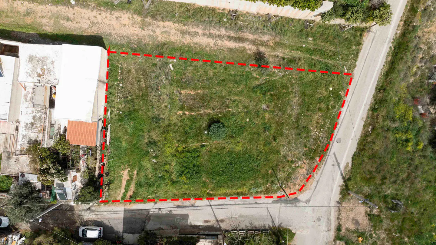 Aerial view of a vacant grassy plot outlined in red, situated next to a white building and a paved road.