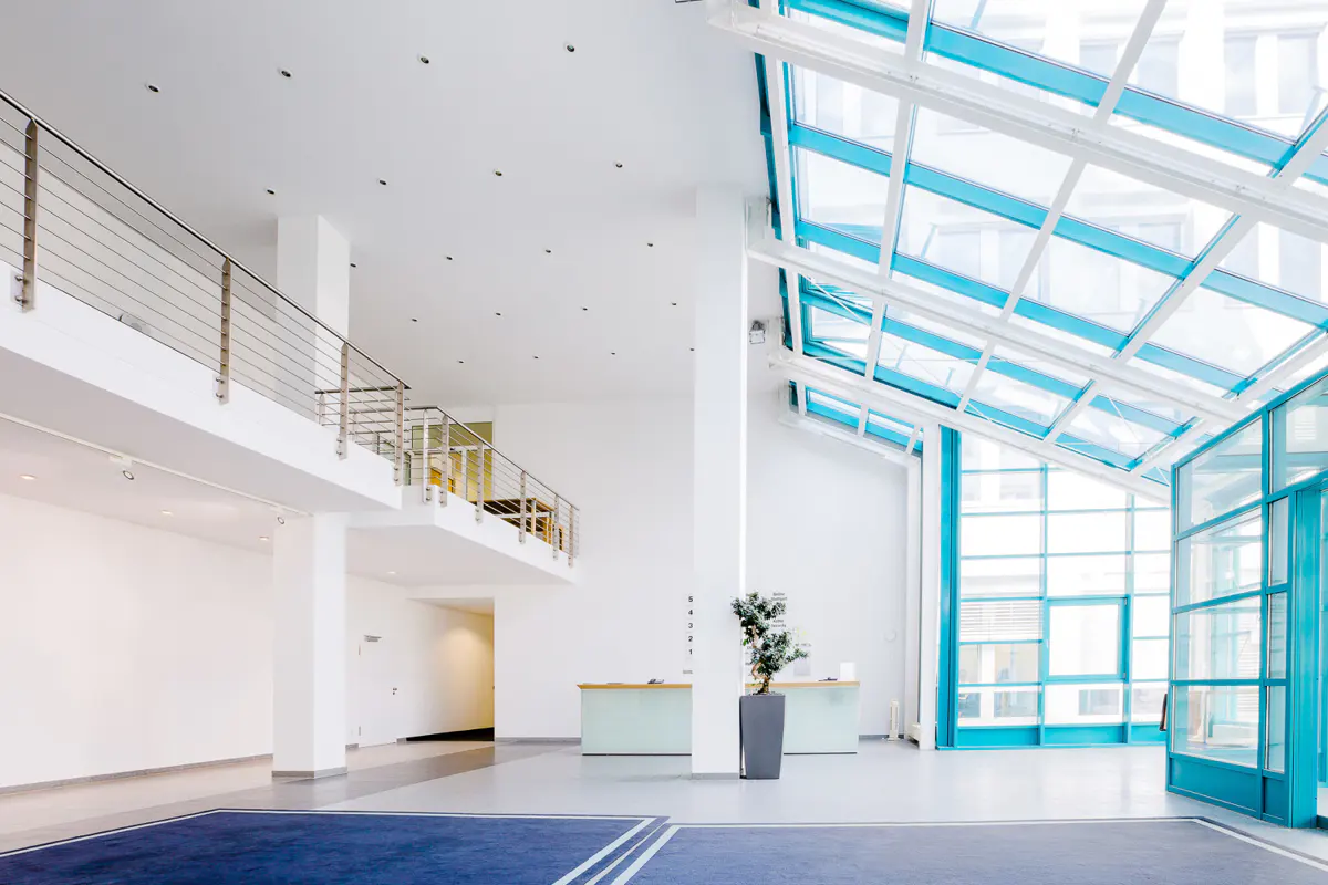 Bright, modern lobby with white walls, blue accents, and a glass ceiling. A second-floor balcony overlooks the space. Reception desks and a potted plant are visible.