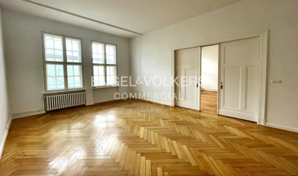 Bright, empty room with herringbone wood floors, white walls, and two large windows. A radiator sits beneath the windows, and sliding doors are partially open.