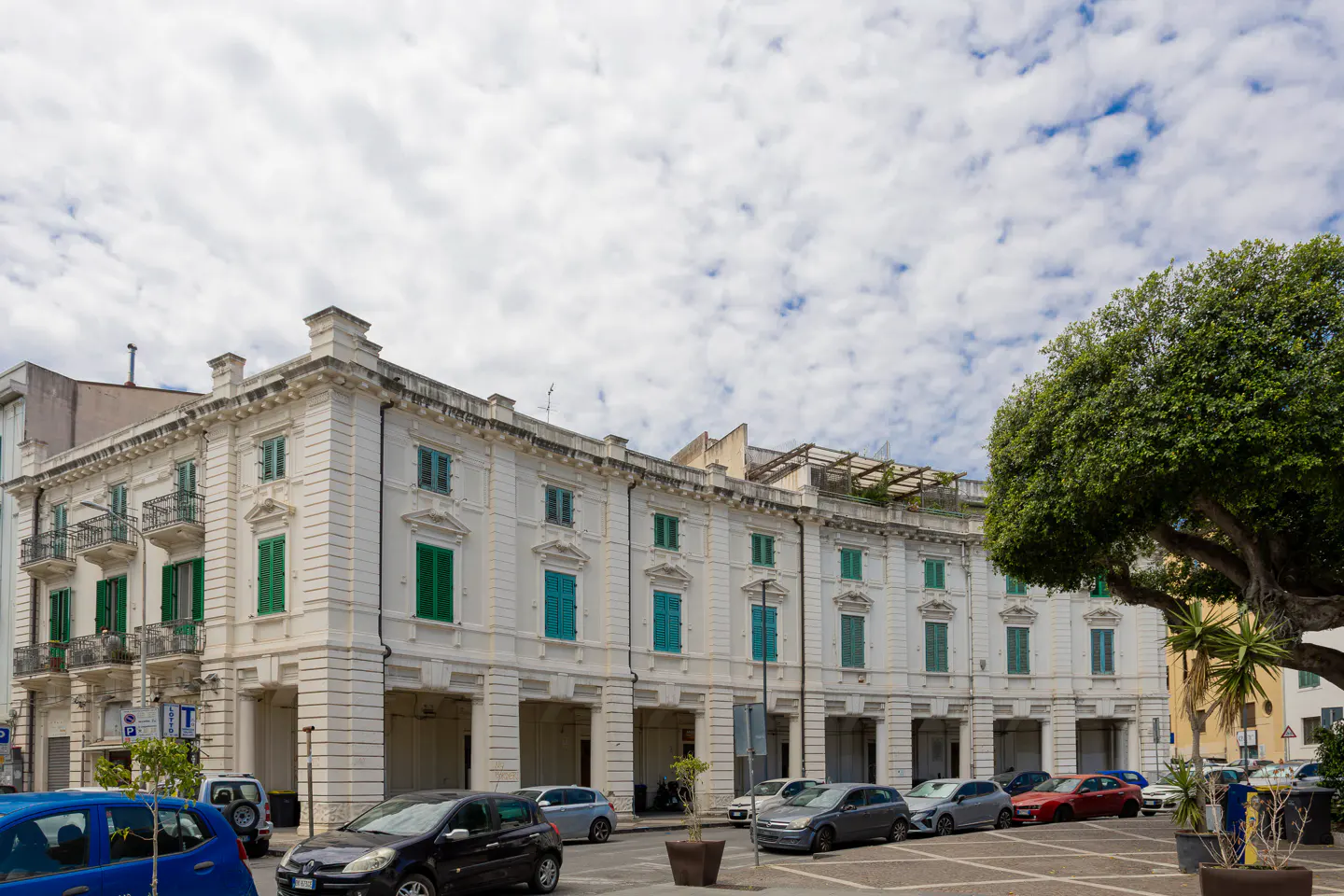 A white historic building with green shutters and arched ground-floor columns, parked cars, and a large tree under a cloudy sky.