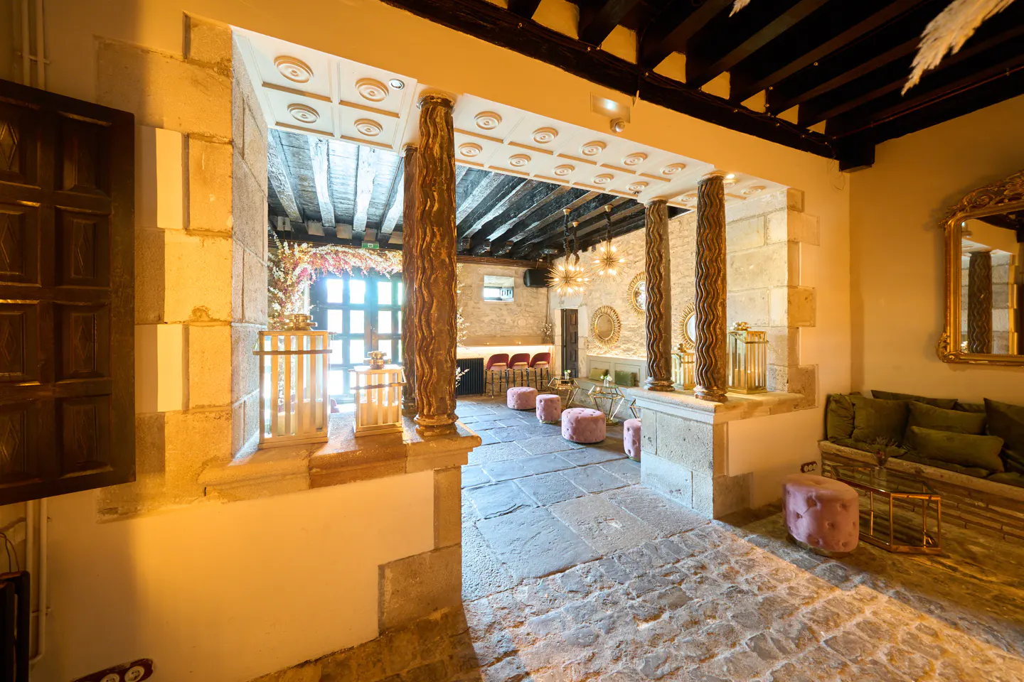 Interior view of a stone building with columns, pink ottomans, and a wooden beamed ceiling.