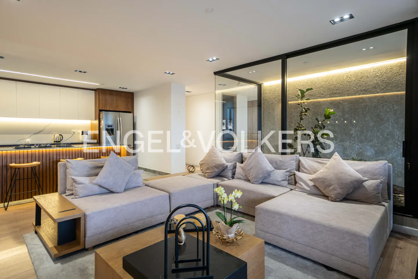 Living room with a large sectional sofa, a modern kitchen, and a glass wall with plants.