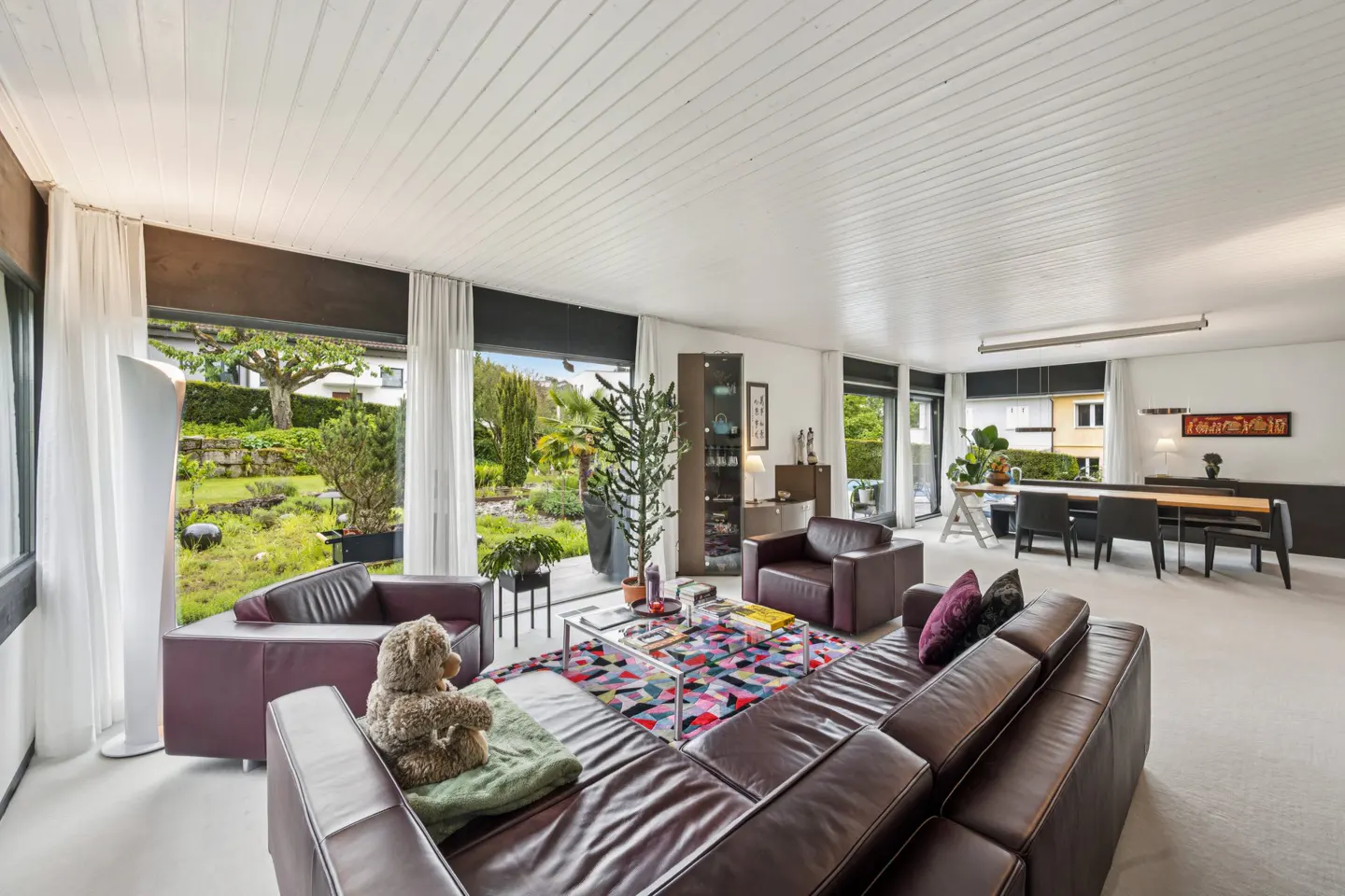 Spacious living room with dark leather sofas, a colorful rug, and large windows overlooking a lush garden.