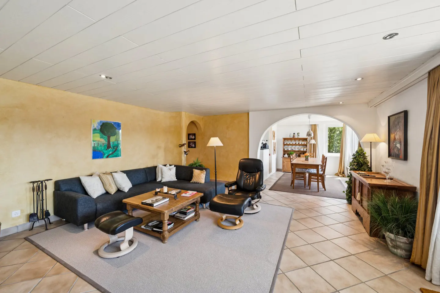 Living room with a gray sectional sofa, leather recliner, and wooden coffee table on a gray rug. Dining room visible through an arched doorway.