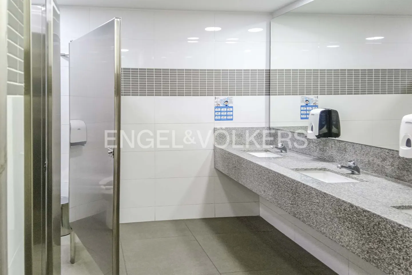 A commercial bathroom with stainless steel stalls, a granite countertop with sinks, and white tiled walls.