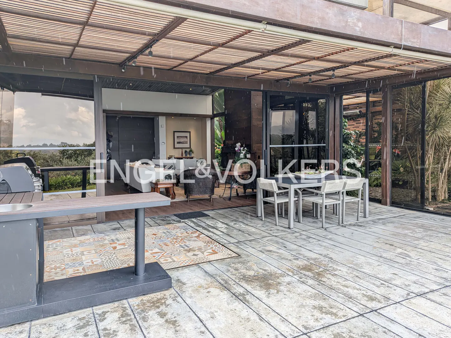 Covered patio with outdoor kitchen, table and chairs. Living room visible through sliding glass doors. It is raining.