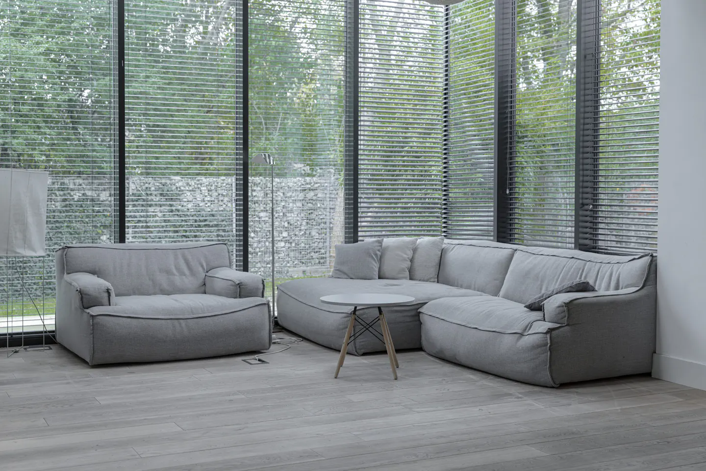 Living room with gray sectional sofa, armchair, and round table. Large windows with blinds overlook a green outdoor space.