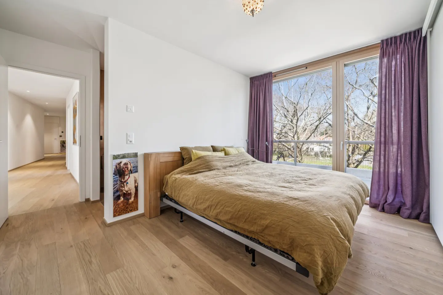 Bedroom with a golden bedspread, wooden floors, and purple curtains by a large window. A hallway is visible through a doorway.