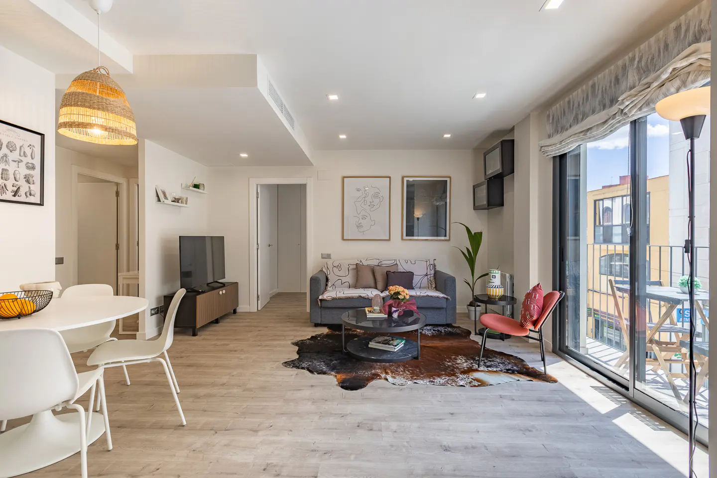 Bright living room with a gray sofa, round table, and a cowhide rug. Balcony doors open to a view of city buildings.