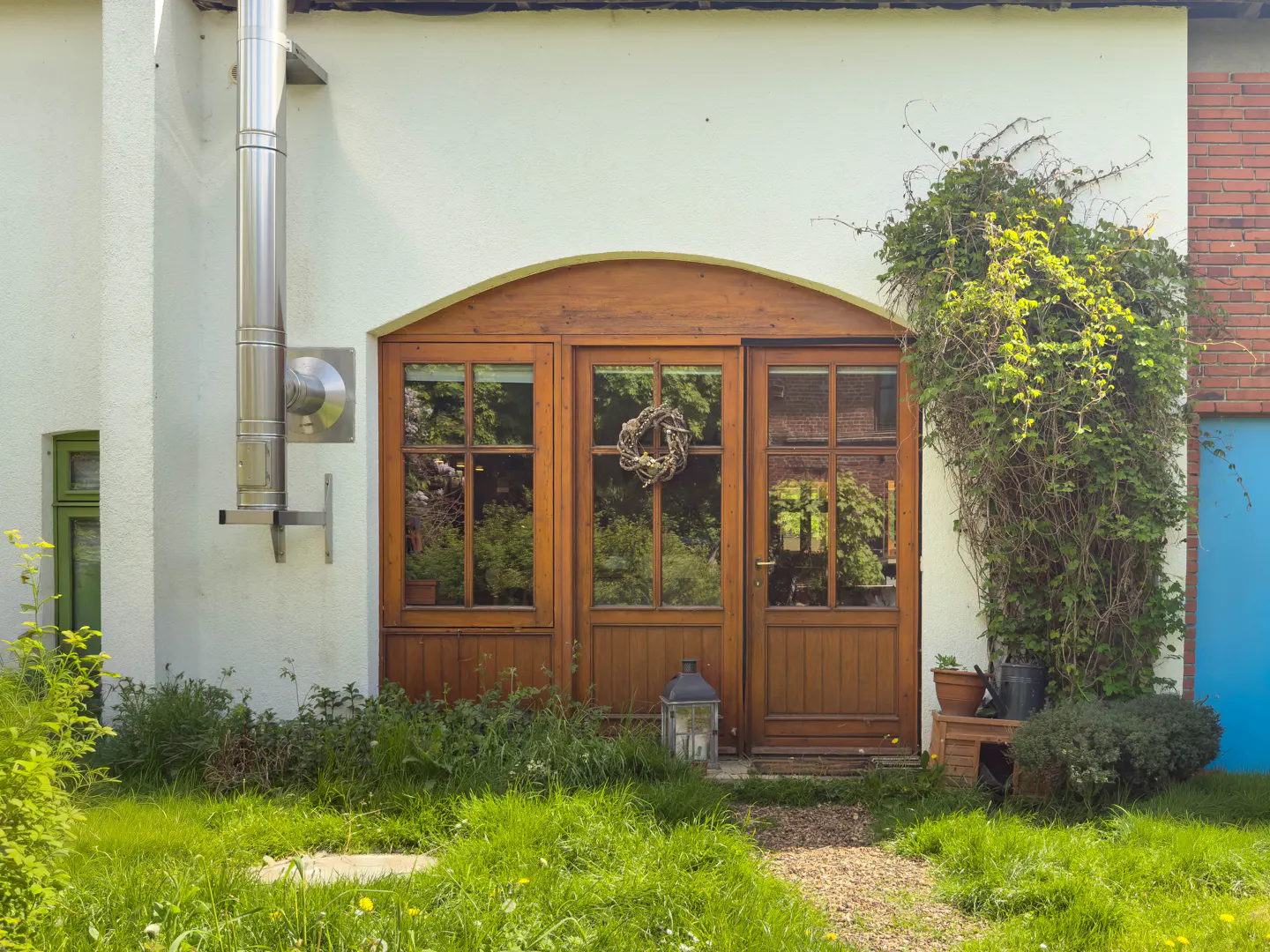 Wooden door with glass panels and a wreath, set in a white building with green grass in front. A metal chimney is on the left.