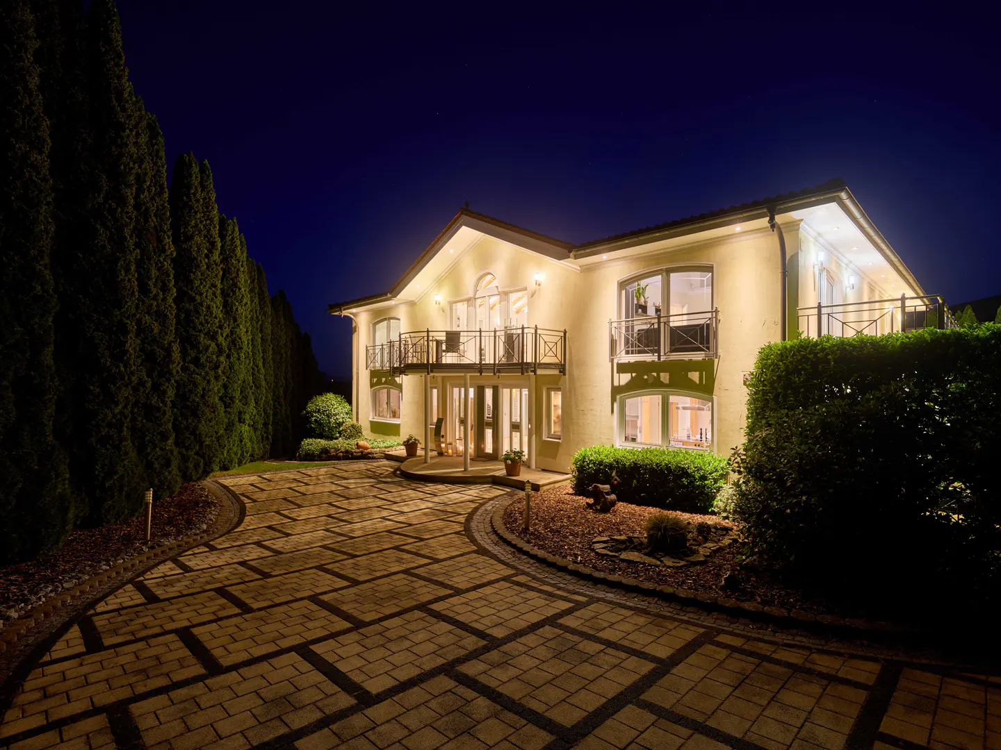 Night view of a two-story cream house with balconies, lit up with warm lights. A patterned brick driveway leads to the entrance, surrounded by green bushes and tall trees.