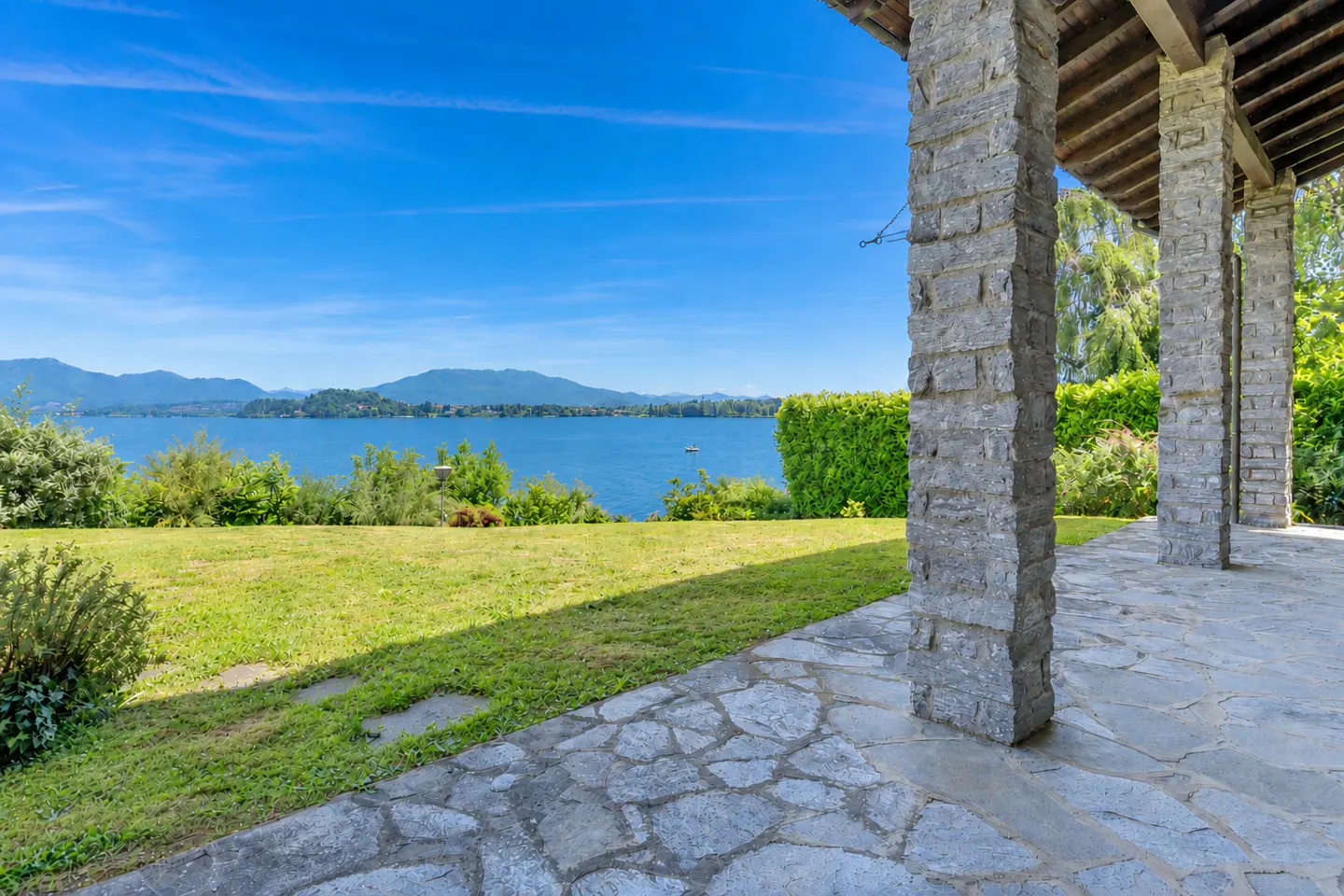 View from a stone patio with pillars overlooking a lake, green lawn, and blue sky.