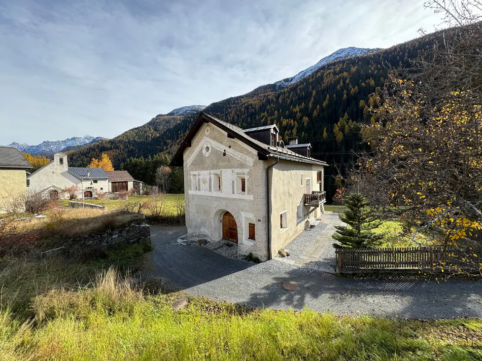 Exterior view of a beige two-story house with a balcony, set against a backdrop of mountains and trees.