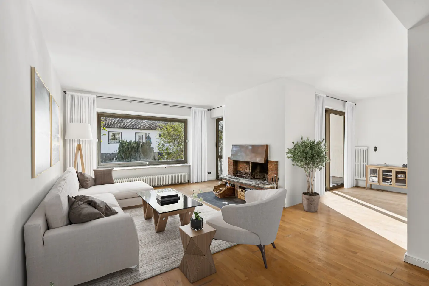 Bright living room with white walls, wood floors, and large window. A white sofa, chair, and fireplace create a cozy atmosphere.