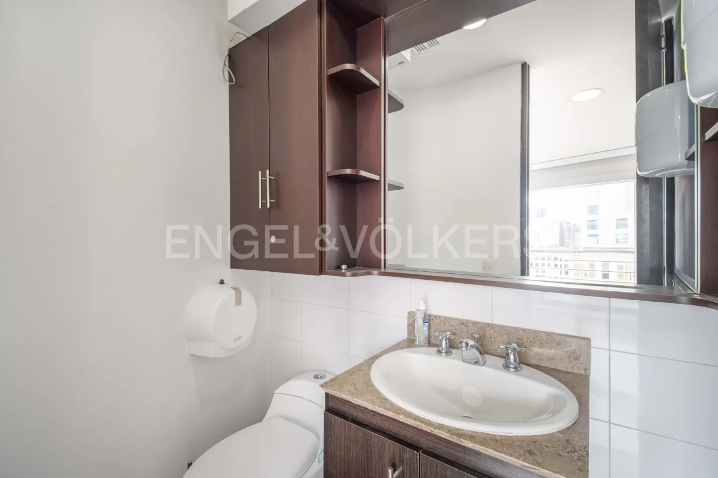 A bathroom with a white toilet, sink, and brown cabinets. A mirror reflects a city view.