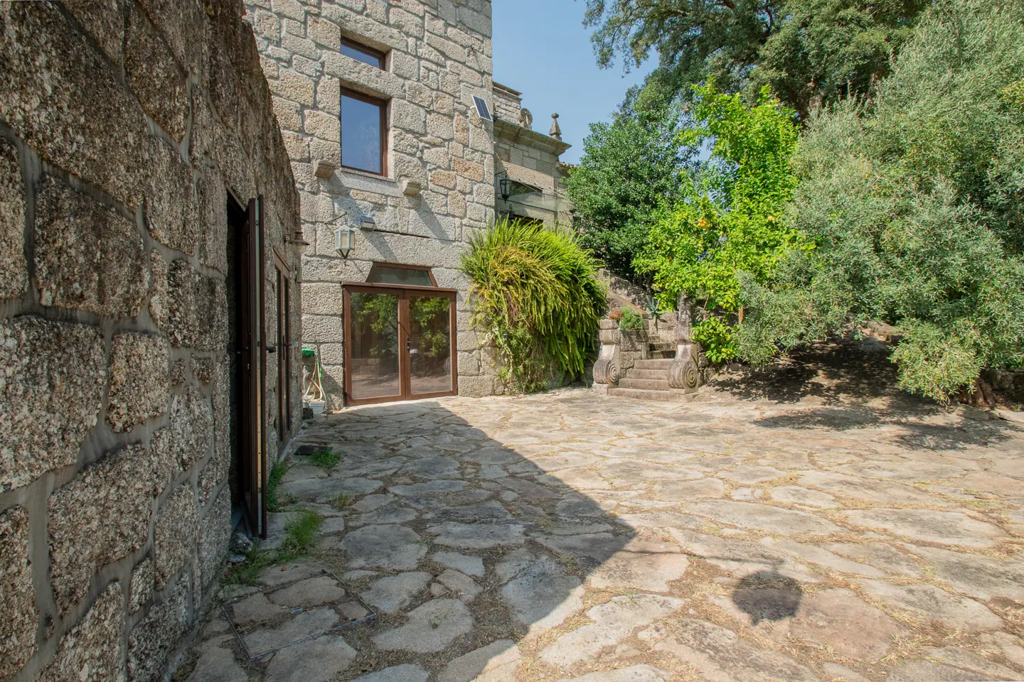A rustic stone house with a flagstone patio, glass doors, and lush green trees under a clear blue sky.