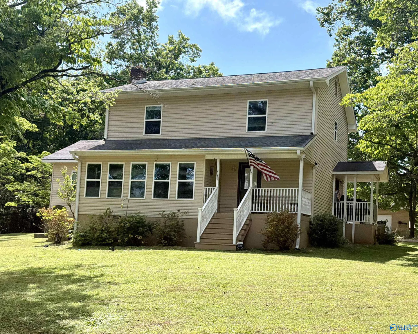 Two-story beige house with a porch, white railings, and an American flag. Green lawn and trees surround the property.