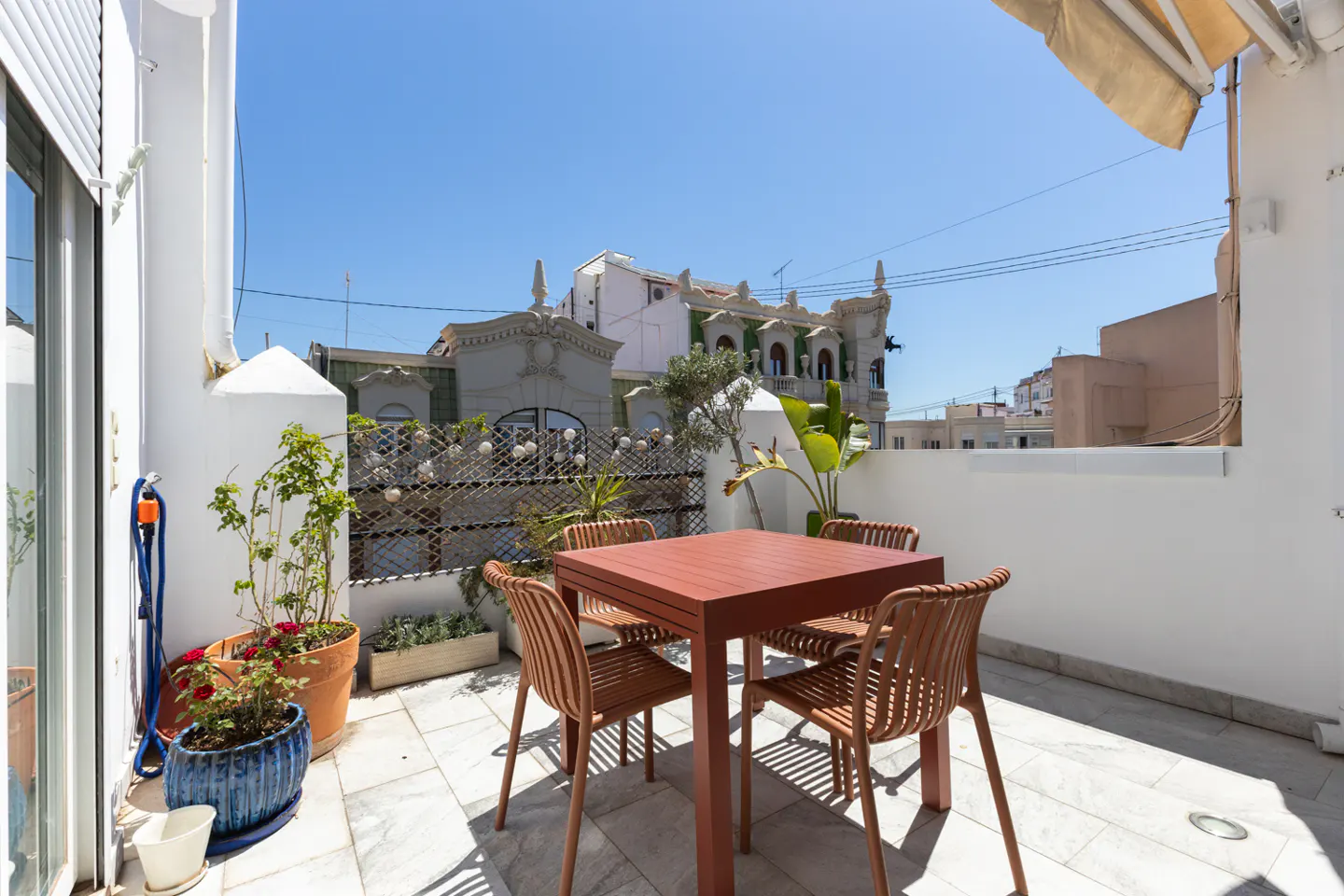 Outdoor patio with a red table and four chairs, potted plants, and a view of buildings under a clear blue sky.