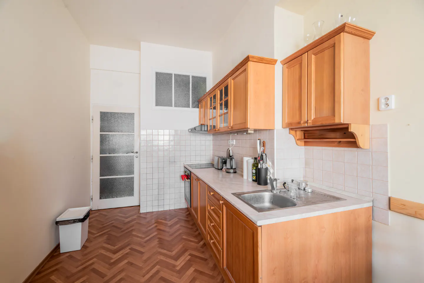Kitchen with light wood cabinets, white tiled backsplash, stainless steel sink, and herringbone wood flooring.
