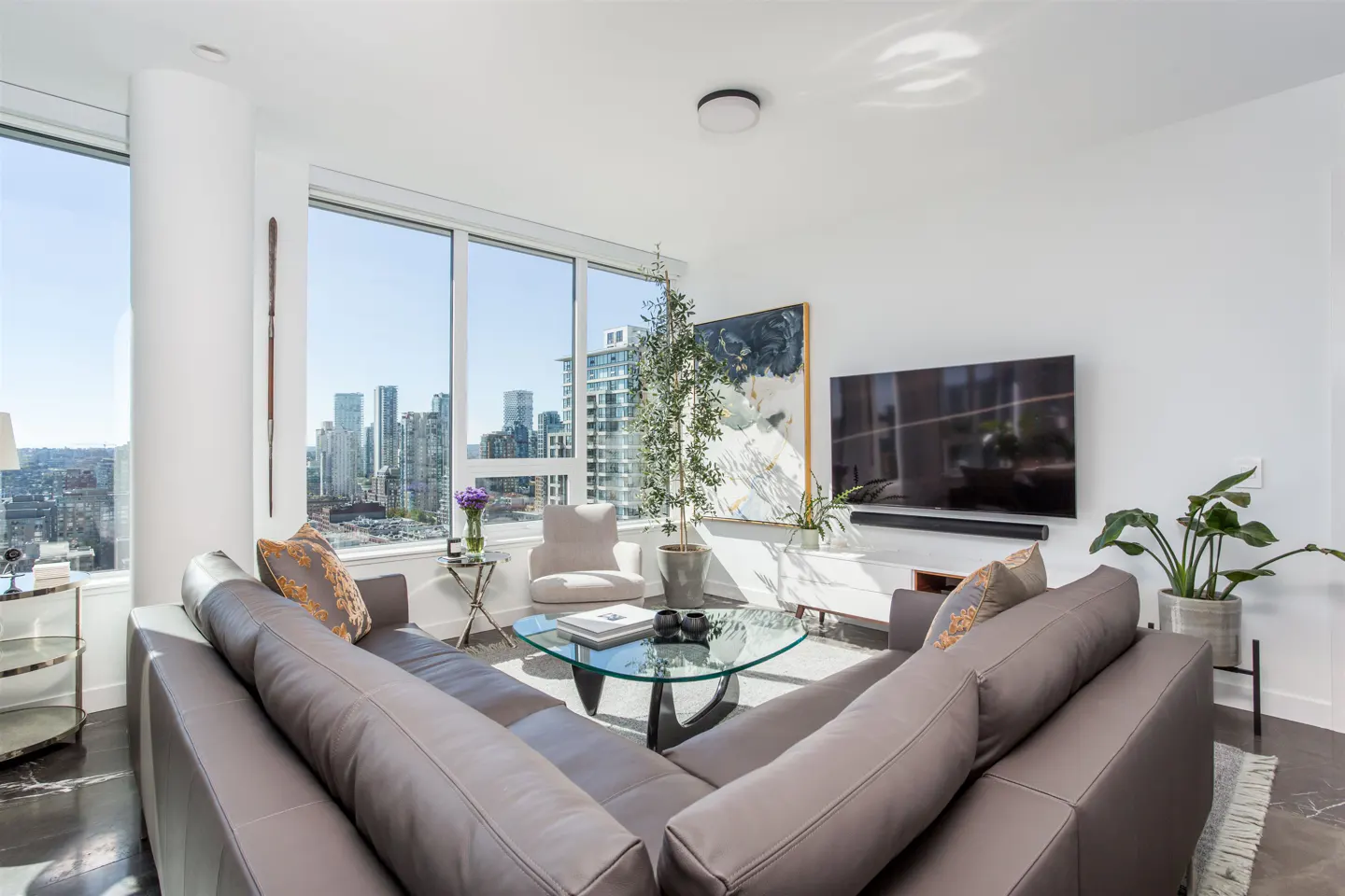 Bright living room with a gray sectional sofa, glass table, and city views from large windows. A TV hangs above a white console.
