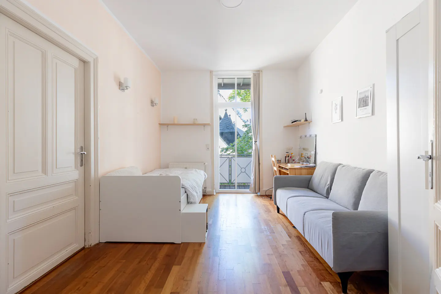 Bright room with hardwood floors, a white bed, a gray sofa, and a desk near a balcony door. Two white doors are on either side of the room.