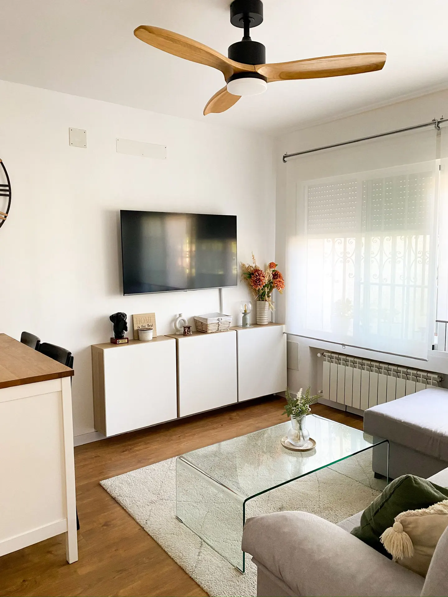 Modern living room with a wall-mounted TV, white floating cabinets, glass coffee table, and a wooden ceiling fan.