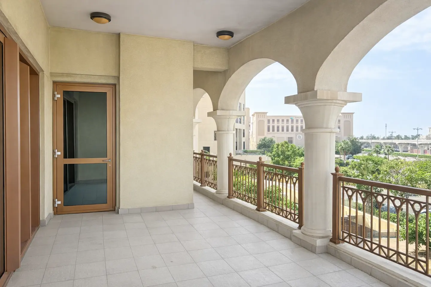 A balcony with white tile flooring, arched columns, and a brown metal railing overlooks green trees and a building in the distance.