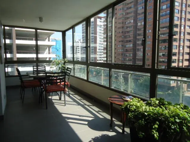 Sunlit balcony with a table, chairs, and plants. Large windows offer a city view with tall buildings.