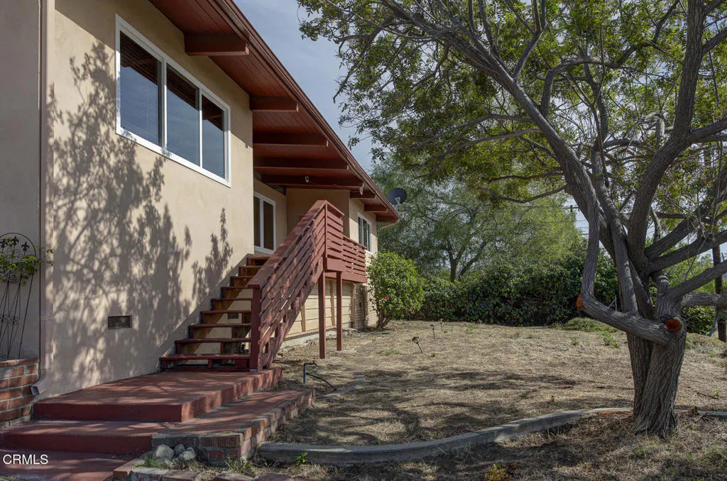 Exterior view of a beige house with red stairs leading to a second-story entrance, surrounded by a dry lawn and a large tree.