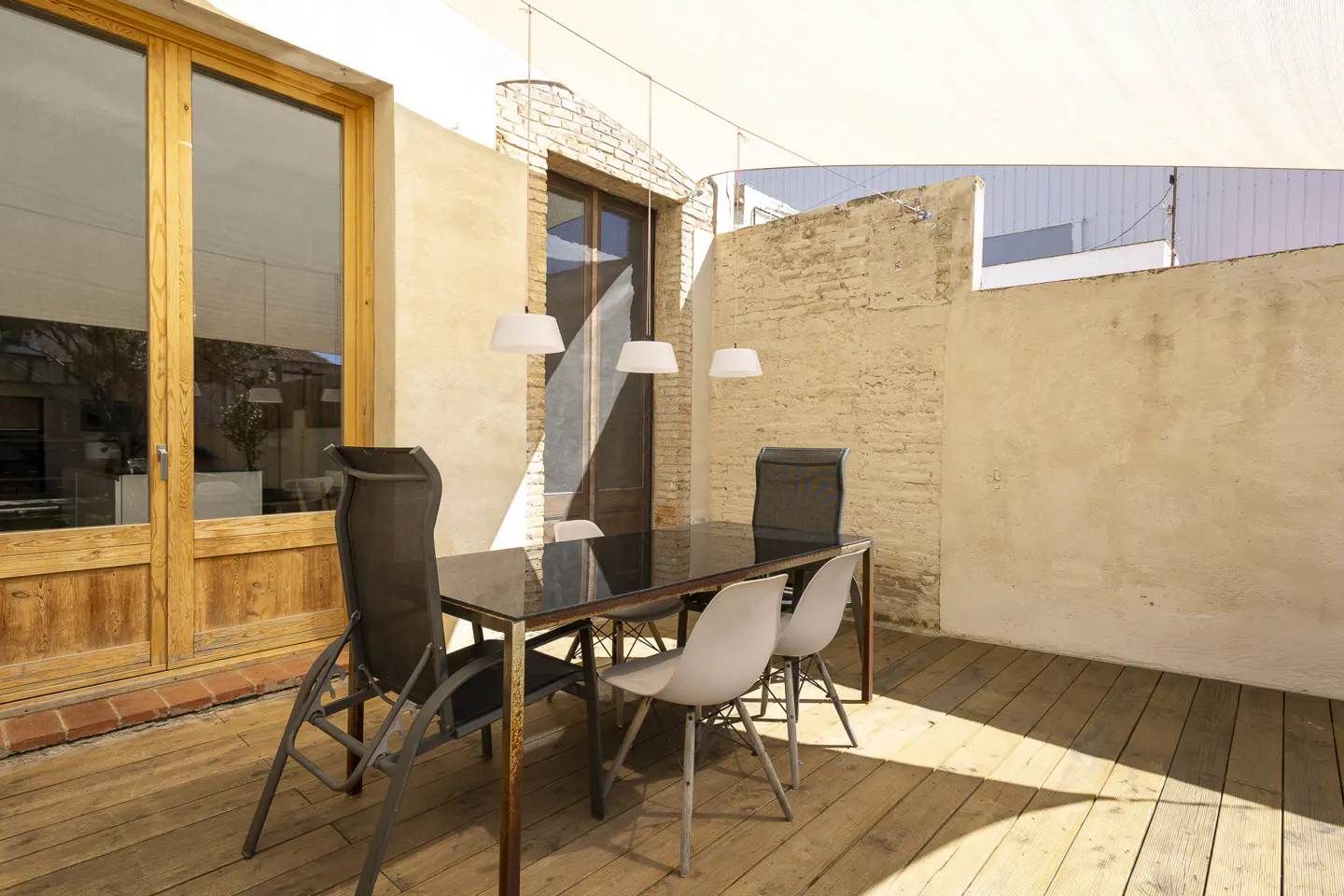 Outdoor dining area with a black table, gray chairs, and wooden deck under a white shade sail. A wooden door and brick wall are in the background.