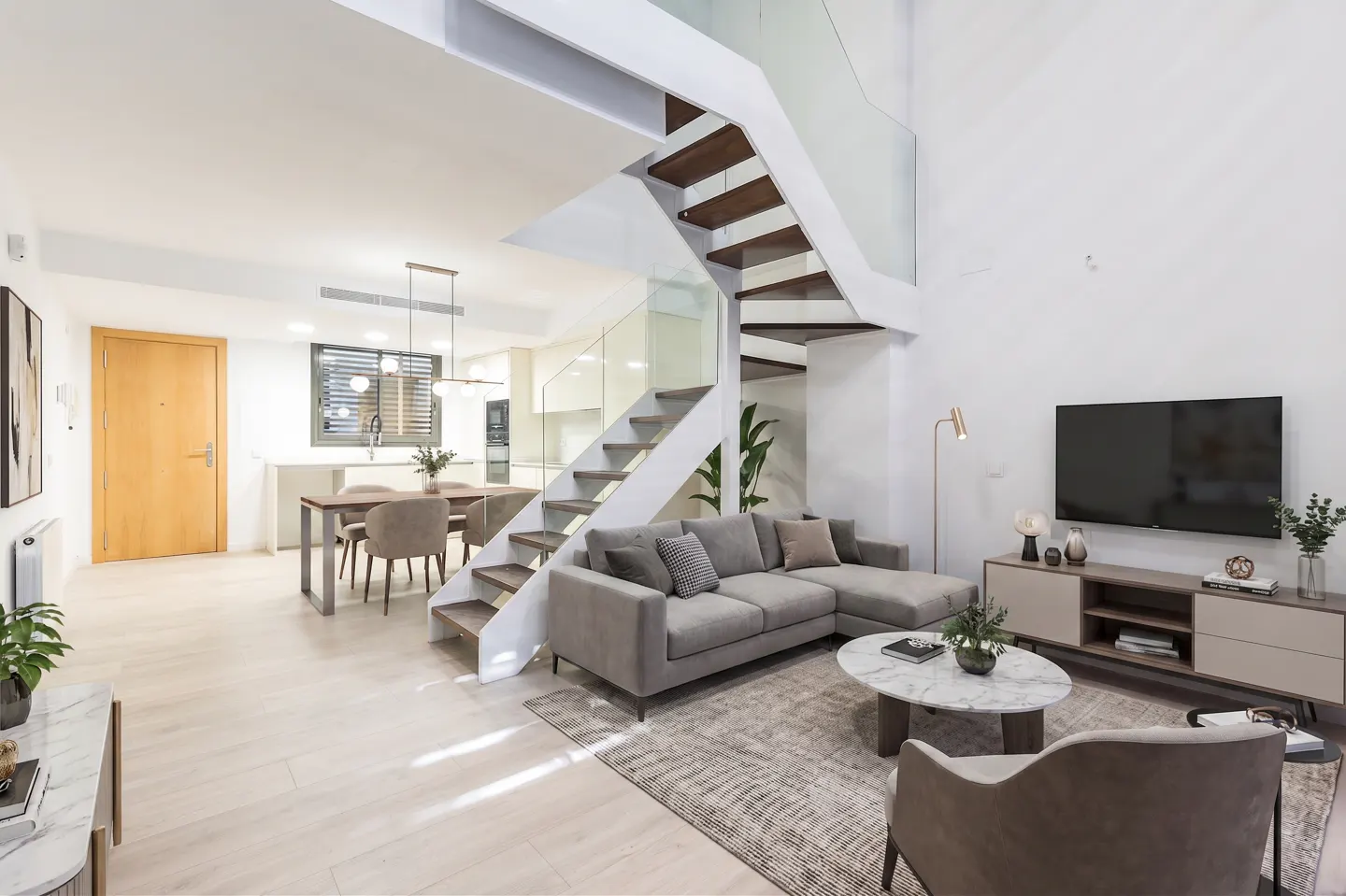 Bright, modern living room with white walls, wood floors, and a staircase with glass railings. A gray sofa and TV stand are visible.