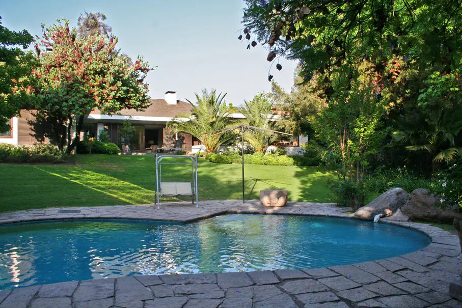 A backyard with a blue pool, stone patio, green lawn, trees, and a house in the background.
