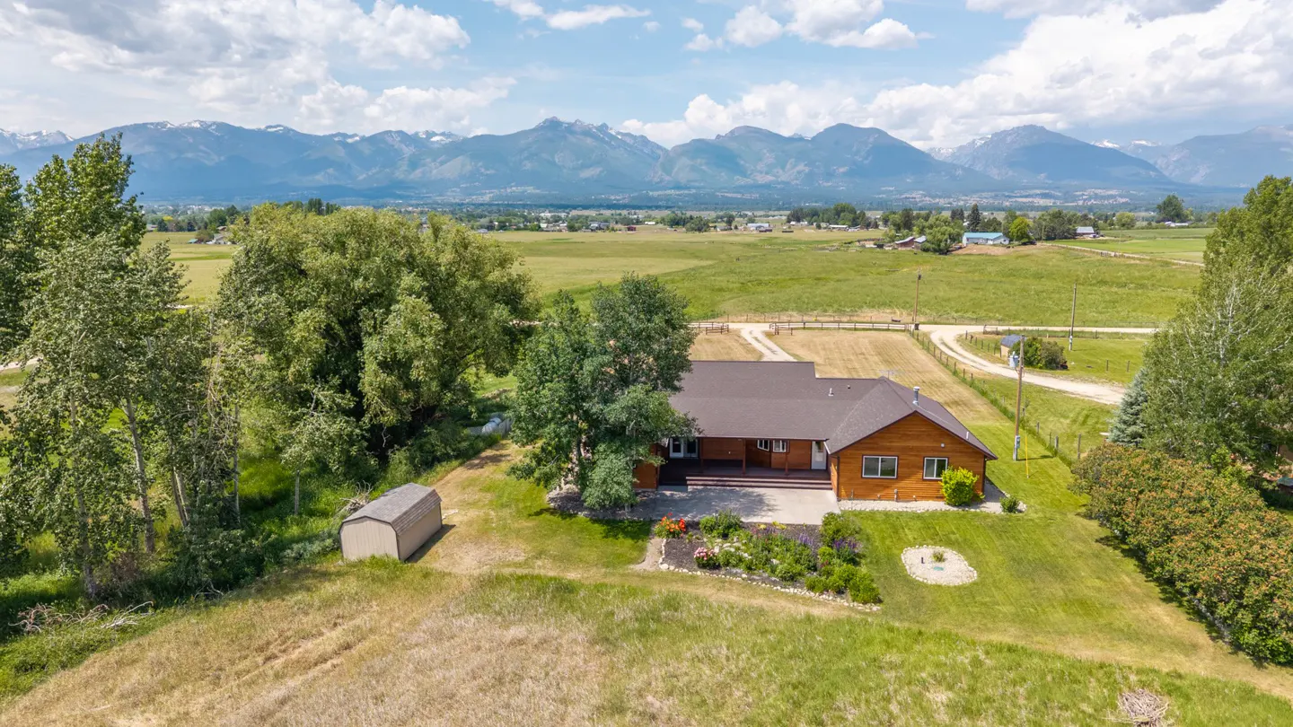 Aerial view of a brown house with a dark roof, green lawn, shed, trees, and mountains in the background.