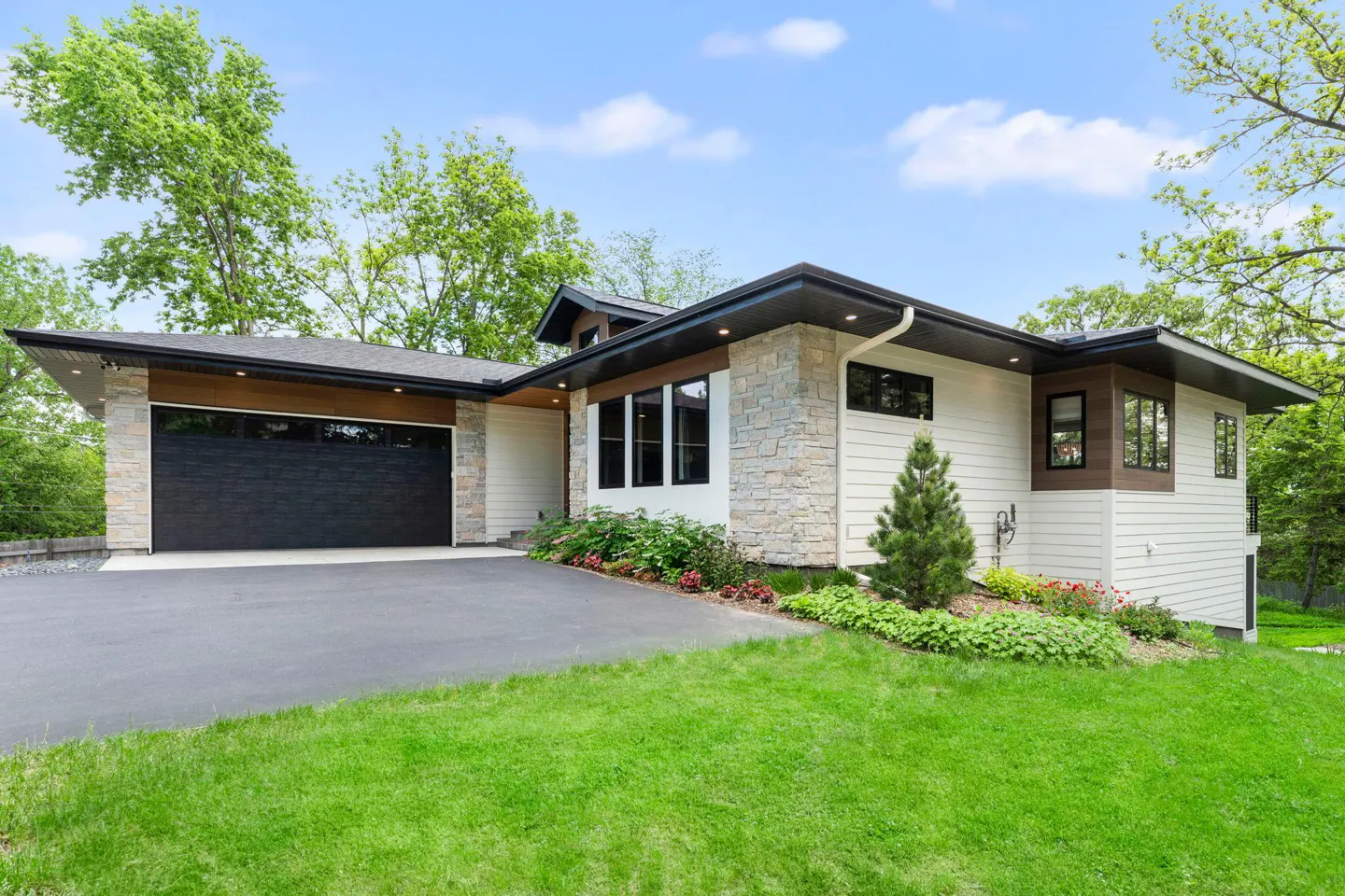 Modern home with a black garage door, stone and white siding, and a green lawn under a blue sky.