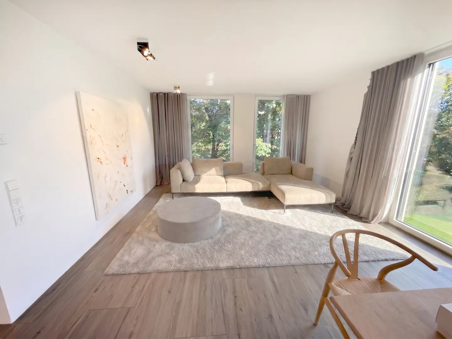 Modern living room with a beige sectional sofa, round ottoman, large abstract art, and floor-to-ceiling windows with trees.