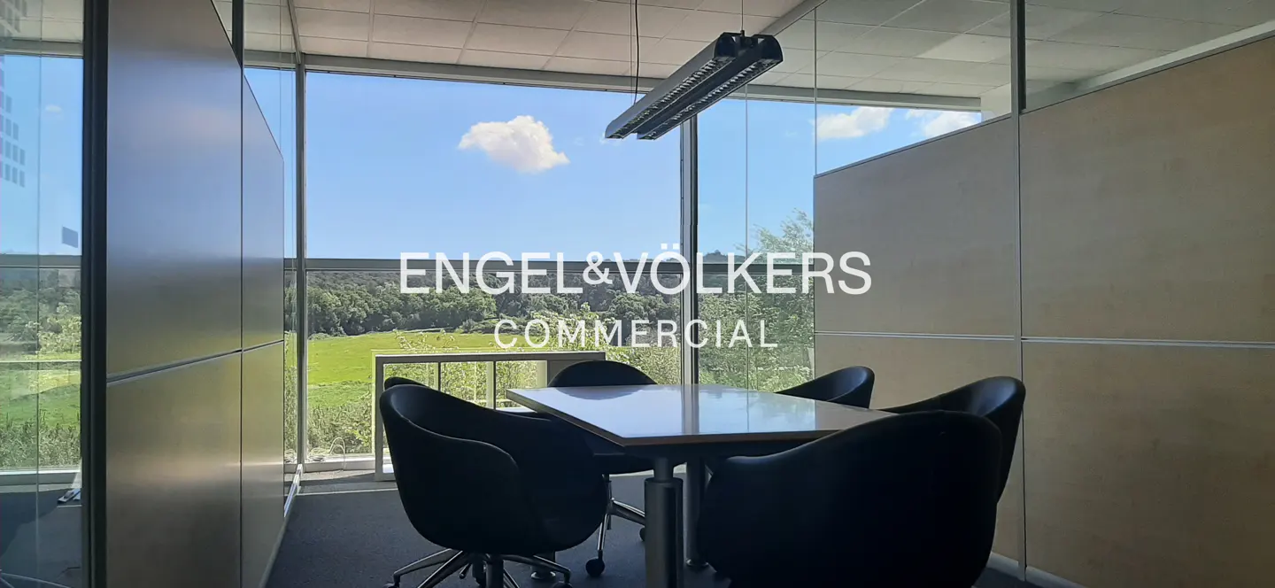 Office meeting room with glass walls, a square table, and four black chairs. Outside, a green field and blue sky are visible.