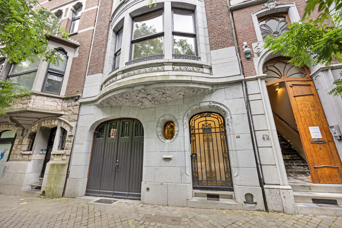 Exterior of a brick building with arched doorways, windows, and a gray stone facade. A wooden door is ajar, revealing stairs.