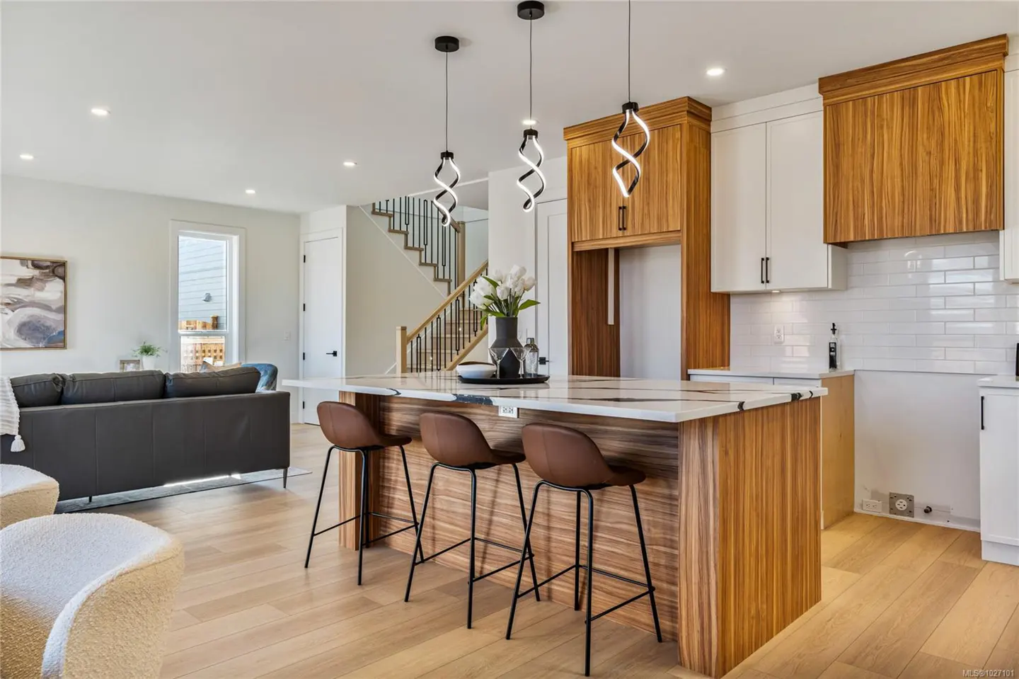 Open concept kitchen with wood cabinets, white countertops, and three brown bar stools. Living room with gray sofa and staircase in background.