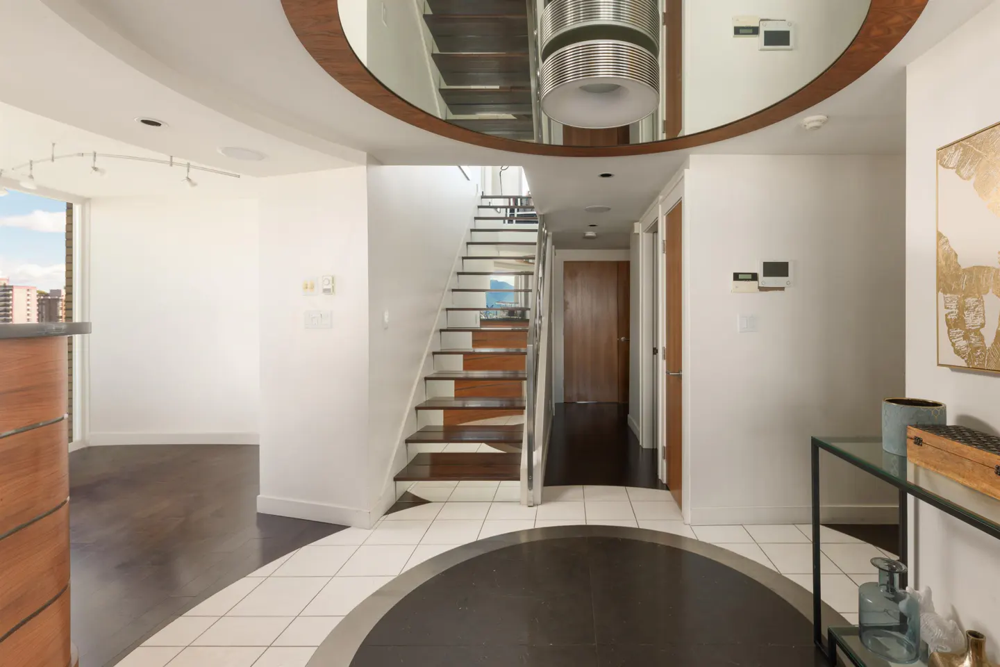 A modern home foyer with white walls, a staircase, and a circular floor design. A glass table and artwork are visible.