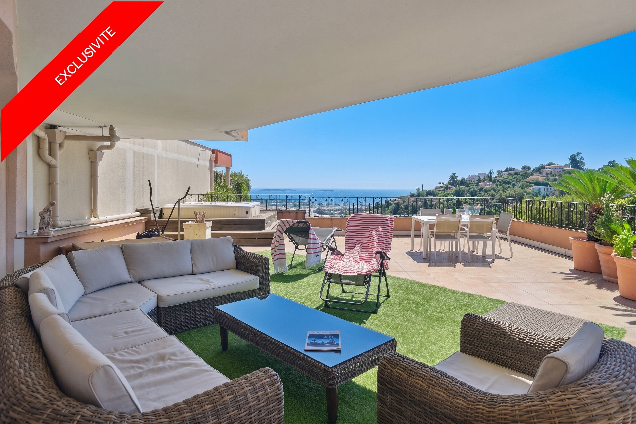 A sunny terrace with wicker furniture, a hot tub, and a dining table overlooks a scenic view of the sea and hills.