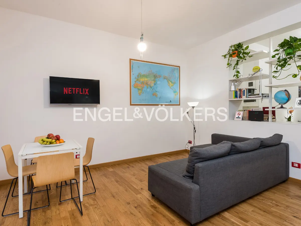 Living room with hardwood floors, gray sofa, white table with fruit, and a Netflix-enabled TV. A world map hangs on the wall.