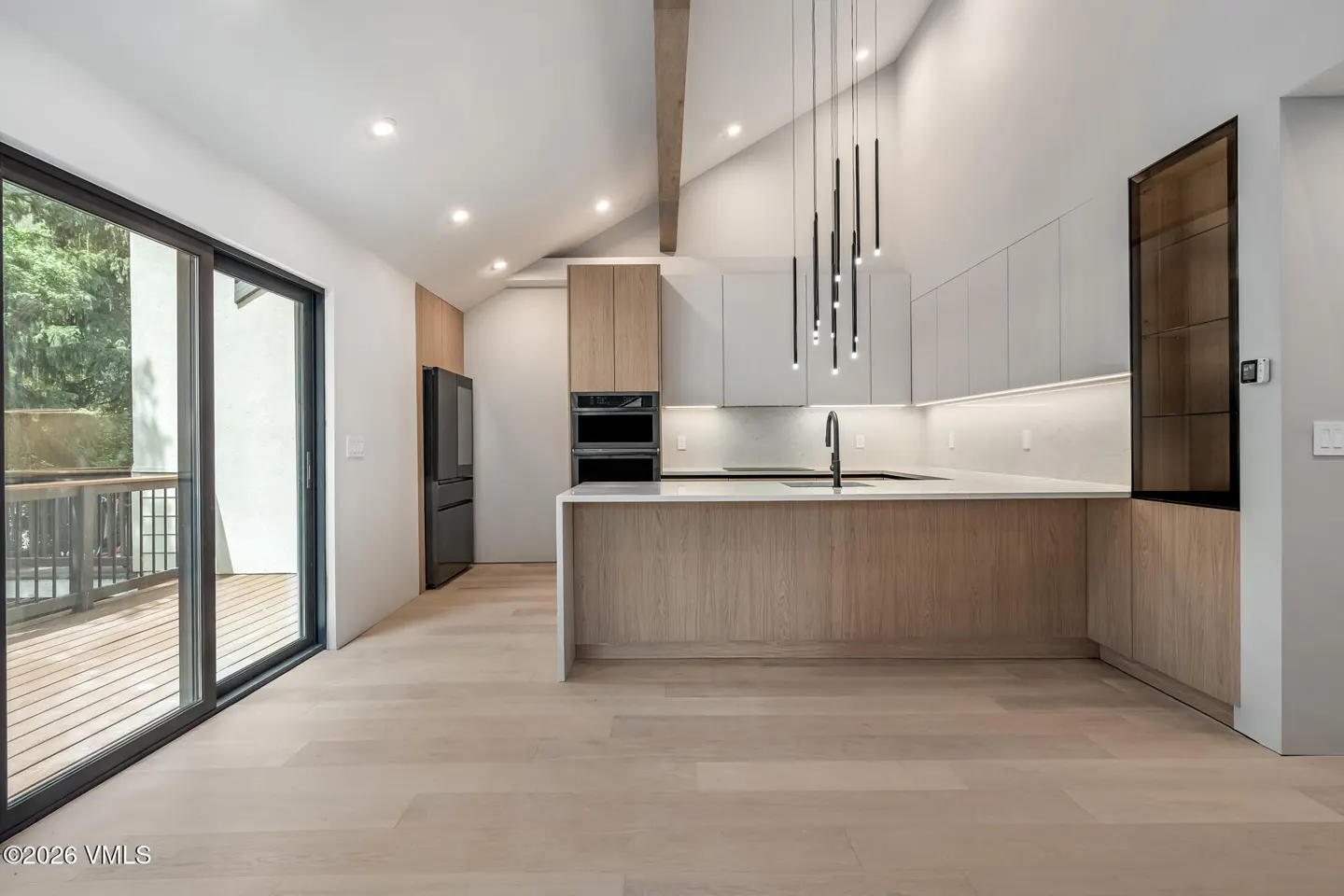 A modern kitchen with light wood floors, a black fridge, and a kitchen island with a white countertop. A sliding glass door leads to a deck.