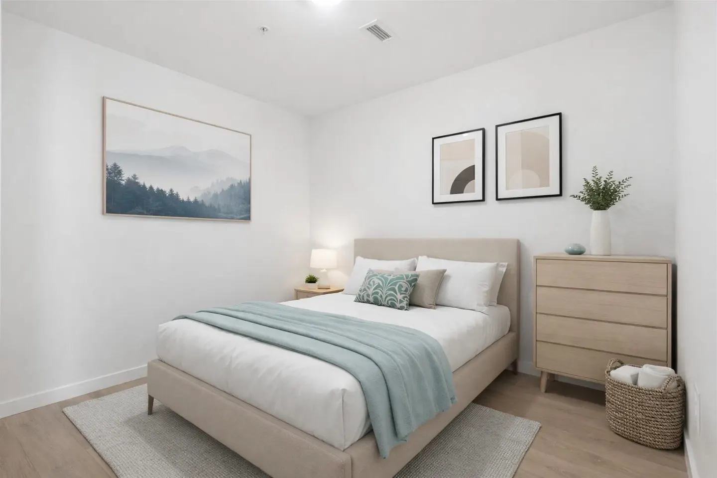 A bright bedroom with white walls, a beige bed with a blue throw, and framed artwork.