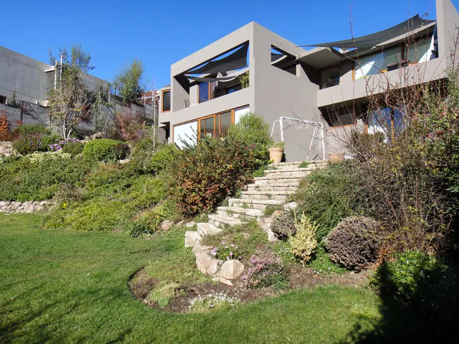 Modern gray house with stone steps leading up to it, surrounded by lush green landscaping and a blue sky.