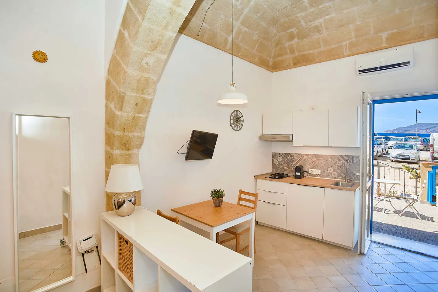 A bright, modern apartment interior with white walls, a stone arch, and a view of parked cars and the sea through an open door.