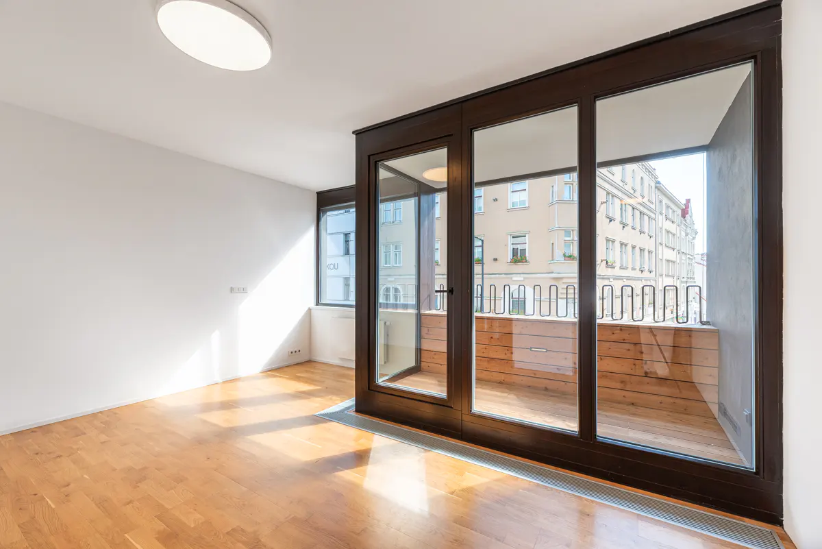Bright, empty room with wood floors, white walls, and a dark-framed glass door to a balcony with a city view.