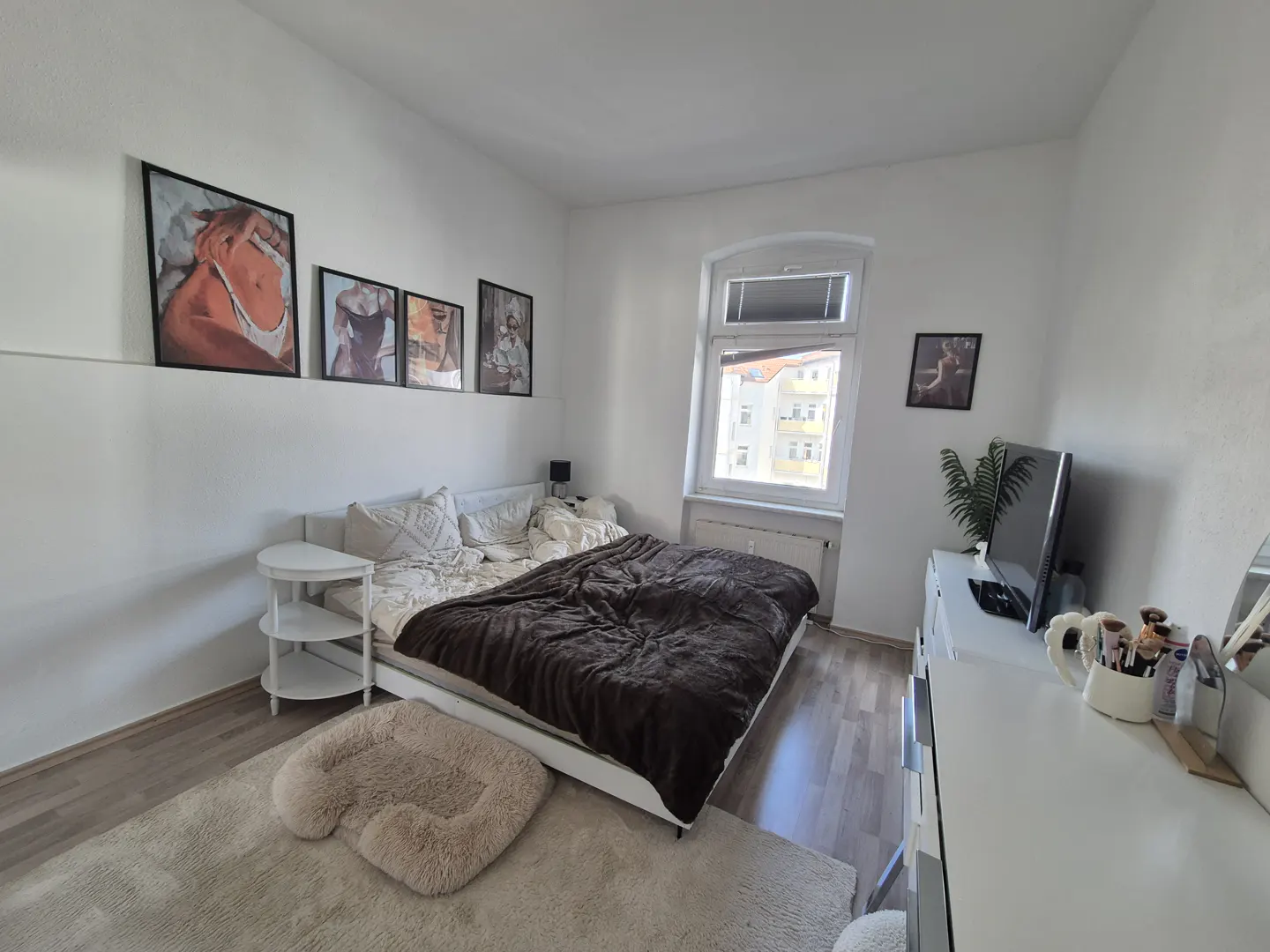 Bedroom with white walls, a bed with a brown blanket, and artwork above the bed. A white dresser and TV are on the right.